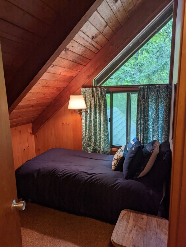 A bedroom with a sloped ceiling, a bed with dark bedding, a nightstand, a lamp, and a window with patterned curtains.
