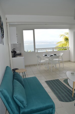 Sala moderna com um sofá azul-turquesa, uma pequena cozinha e uma mesa de jantar com cadeiras. Há uma janela grande com vista para o mar e palmeiras.