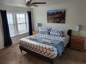 A bedroom with a bed, two bedside tables, a ceiling fan, a window with curtains, and a wall-mounted picture. - Spacious 3 BDR 2.5 BA House with Hot tub, BBQ, Fire Pit, Night View, Playground (Colorado Springs)