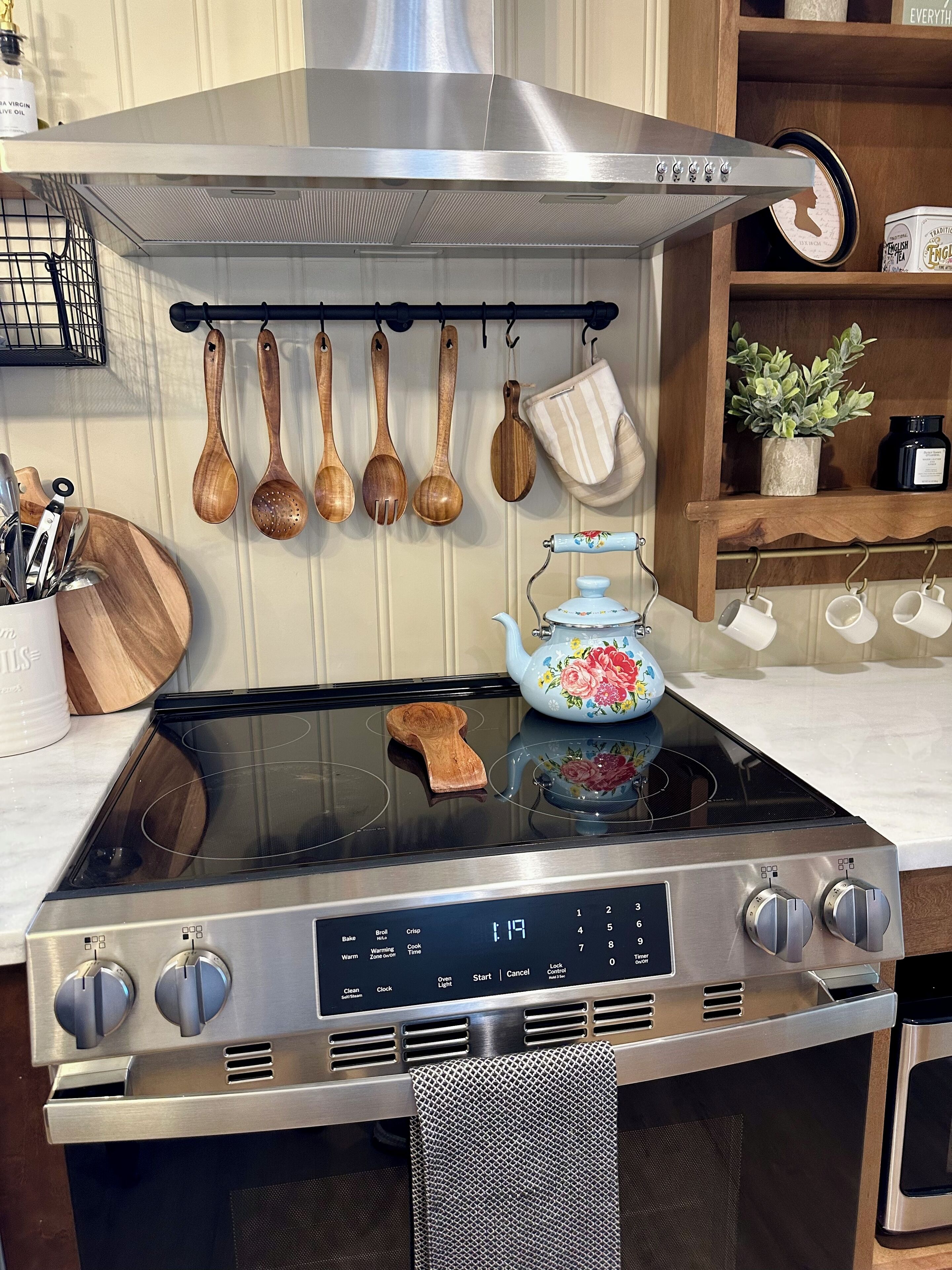 Cucina con piano cottura in acciaio inox, utensili in legno, bollitore e ripiano con tè e una pianta.