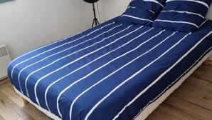 Un lit recouvert d’une couvre-lit à rayures bleues et blanches, dans une pièce dont le sol est en bois et où se trouve un lampadaire noir.
