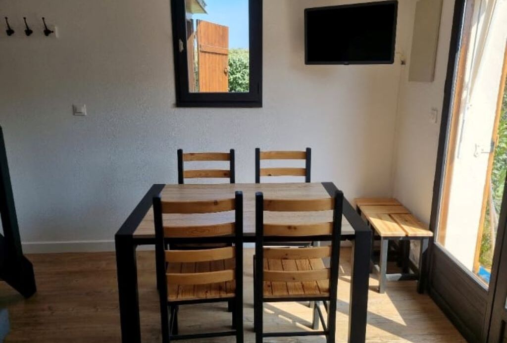 Un comedor con una mesa y cuatro sillas, un pequeño banco, un ventanal con vistas a zonas verdes y un televisor montado.