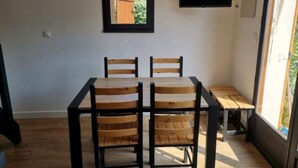 Un coin repas comprenant une table et quatre chaises, un petit banc, une fenêtre donnant sur un espace verdoyant et un téléviseur fixé au mur.