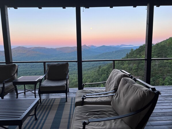 A balcony with a view of mountains and a sunset. There are two chairs and a table on the balcony.