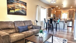 A living room with a brown leather sofa, a coffee table, and a painting of a cityscape. - Charming 2BR Cottage • Near Freeway-Cul-de-sac (Medford)