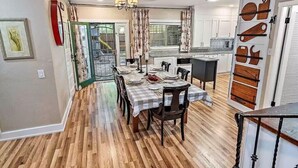 A dining area with a checkered tablecloth, wooden flooring, and a kitchen with white cabinets and a backsplash.