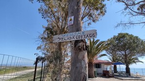 A wooden signpost with 'WELCOME DELADA COTTAGE' written on it, a small building with a red roof, and a beach area with a fence and palm trees. - Beachfront/2br/garden/bbq/seaview/quietretreat (Mesolongi)