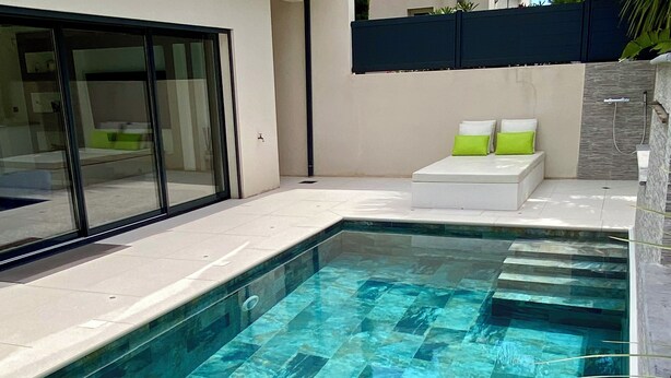 Una piscina rectangular con un diseño de mosaicos, un área de descanso blanca con cojines, y una puerta de cristal que da a una habitación.