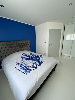 Ein Schlafzimmer mit einem Bett, einem gesteppten Kopfteil, einer blauen Akzentwand, einer weißen Tür und einem Glastisch.