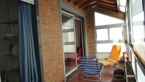 Un balcon avec un mur en briques, une porte-fenêtre coulissante, une chaise à rayures bleues, une chaise orange et une petite table sur laquelle est posée une plante en pot.