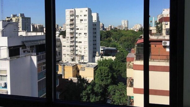 Une vue à travers une fenêtre sur un paysage urbain, avec de hauts immeubles, dont un bâtiment blanc imposant doté de nombreux balcons.