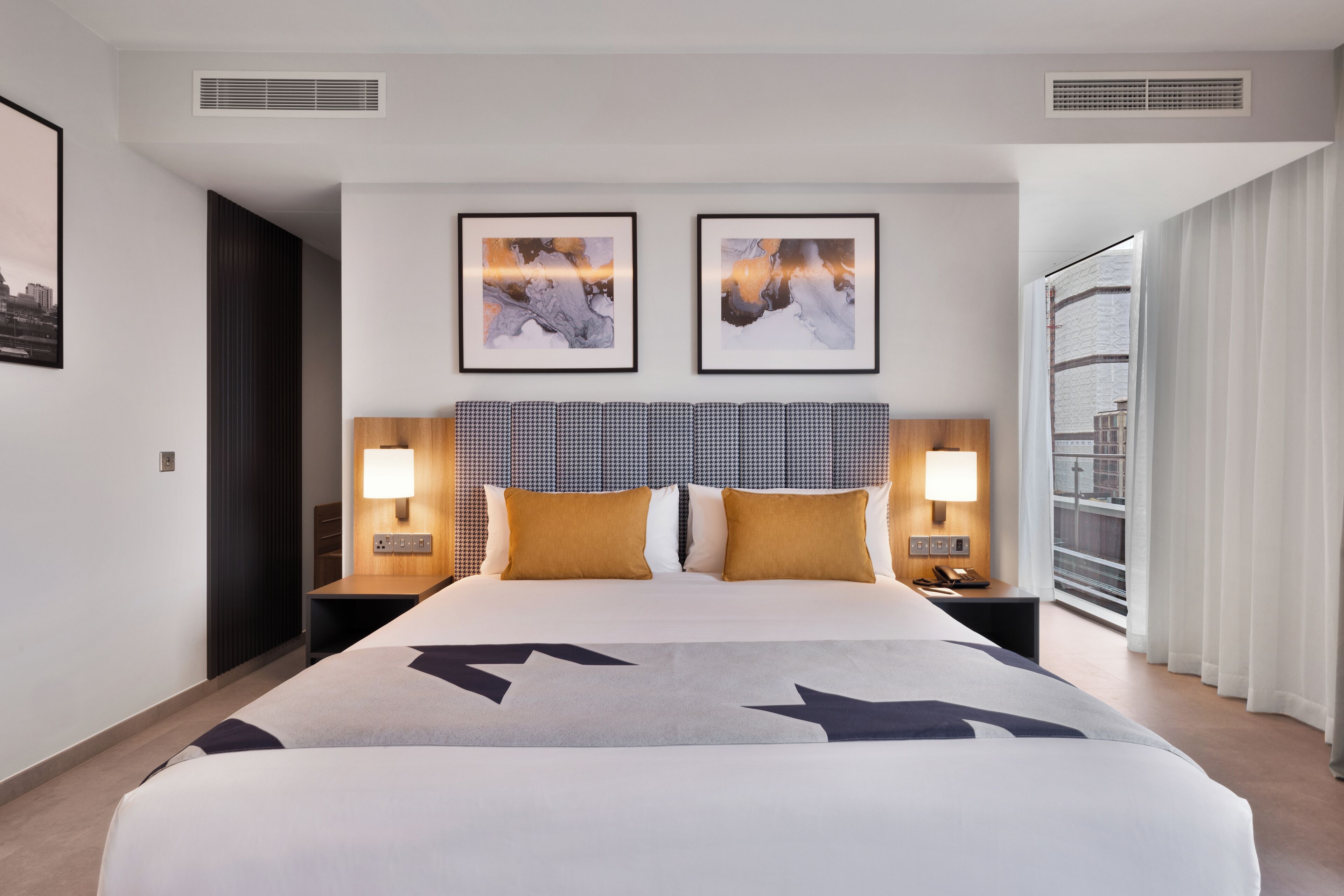 A hotel room with a large bed, two bedside lamps, a headboard with a checkered pattern, and two framed pictures on the wall.
