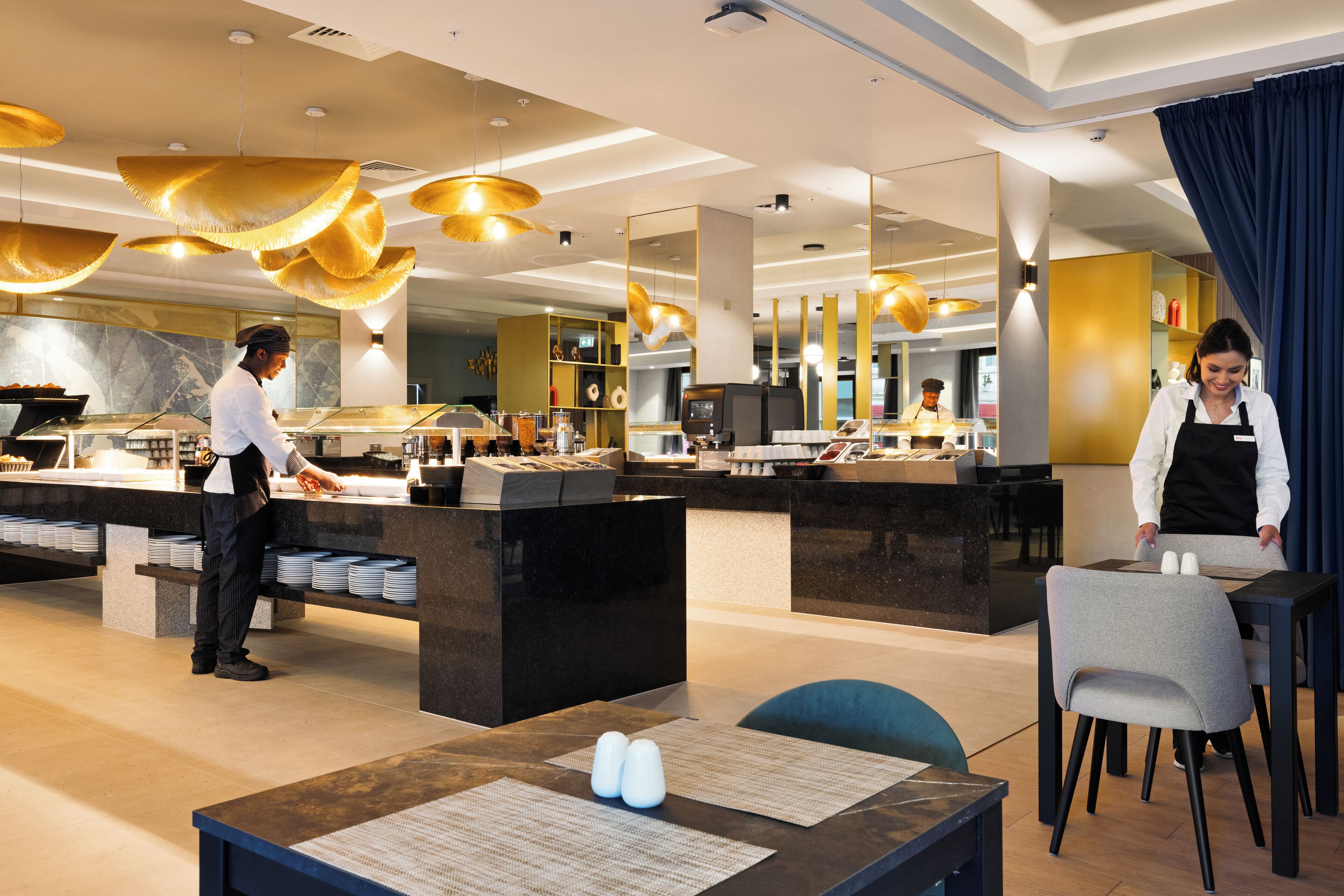 Un restaurante de hotel moderno con un chef preparando comida, un camarero sirviendo y un comedor con mesas y sillas.