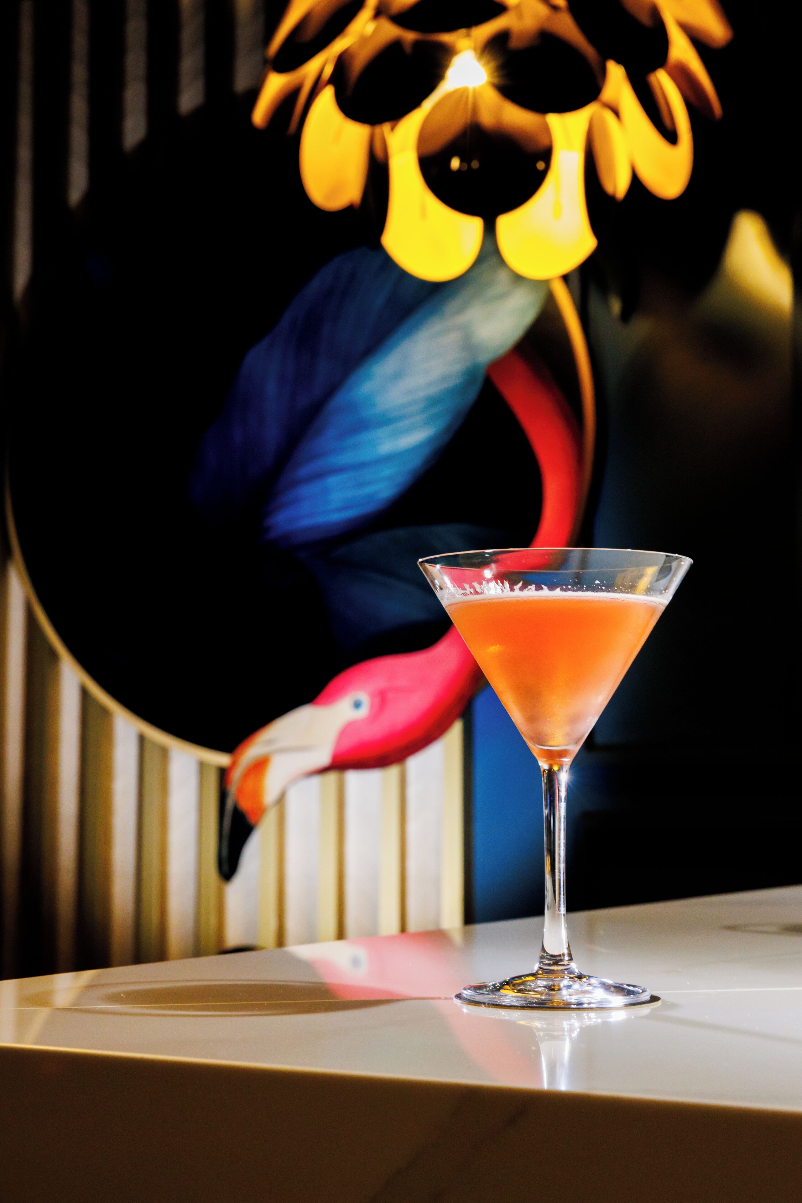 A martini glass with a clear liquid on a reflective surface, set against a backdrop with a colorful illustration of a flamingo.