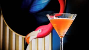 A martini glass with a clear liquid on a reflective surface, set against a backdrop with a colorful illustration of a flamingo.