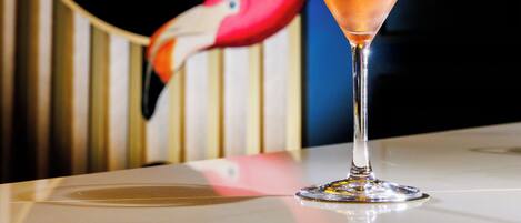 A martini glass with a clear liquid on a reflective surface, set against a backdrop with a colorful illustration of a flamingo.