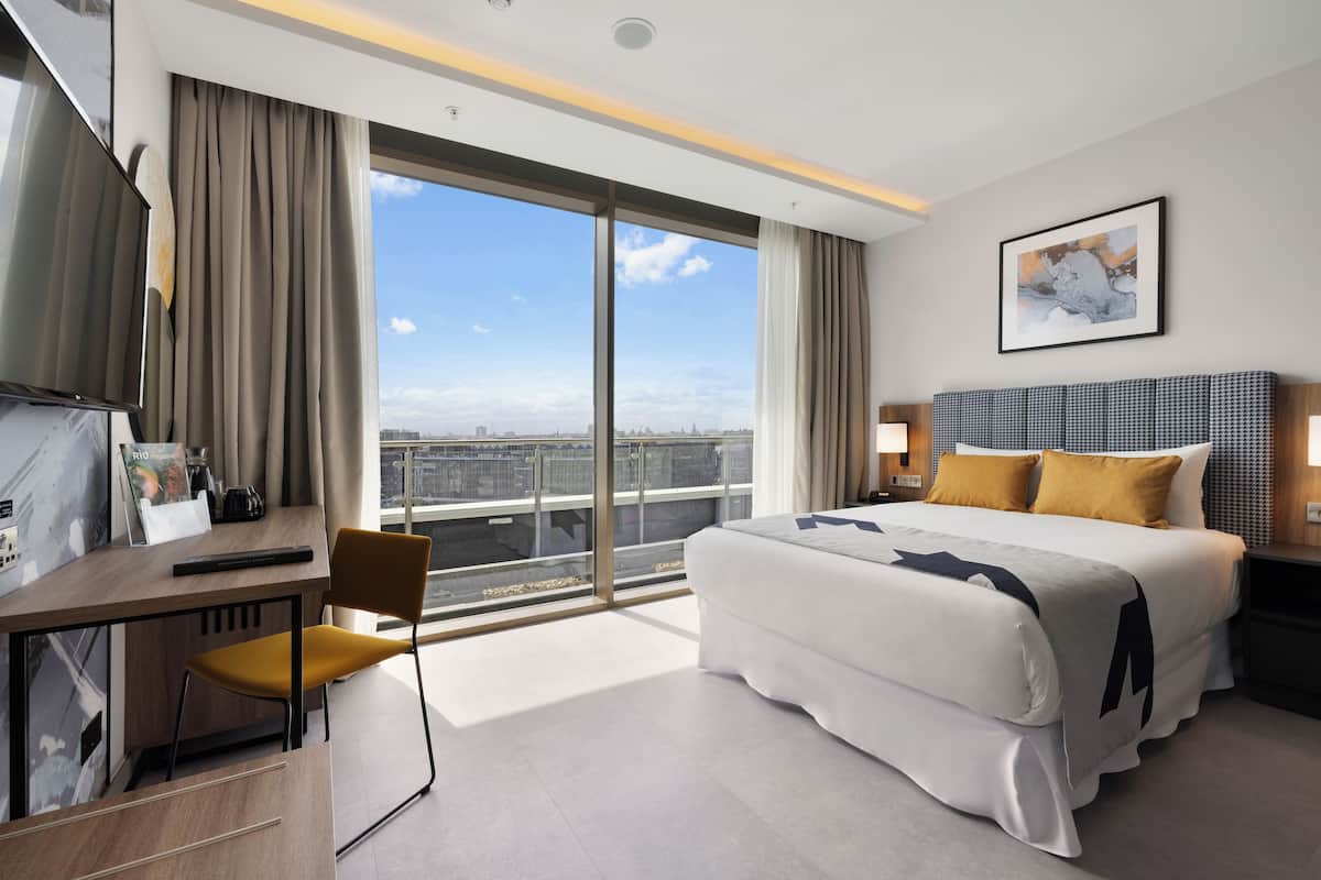 A modern hotel room with a large bed, a desk with a chair, a TV, and a city view.