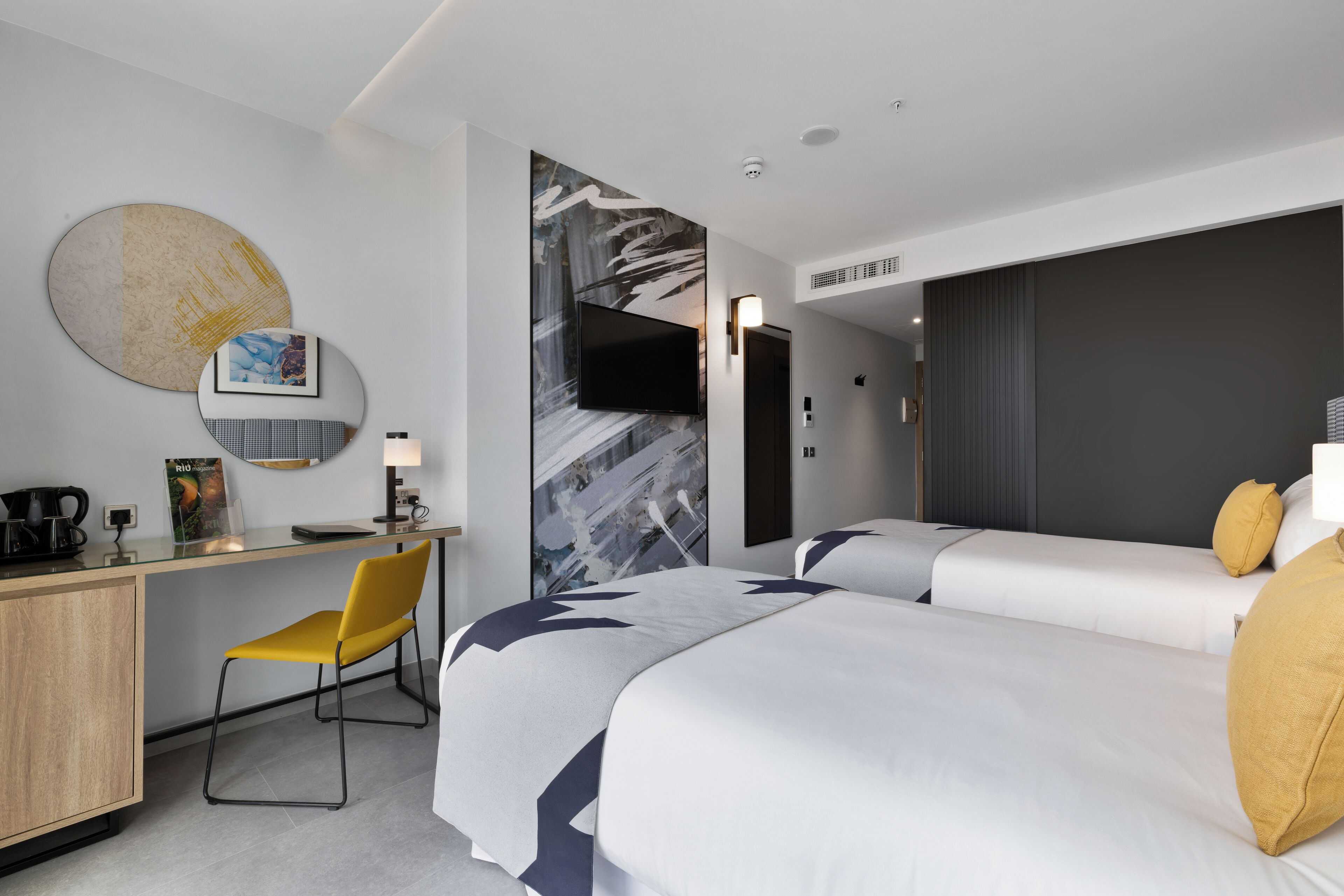 A modern hotel room with two beds, a desk, and a chair.