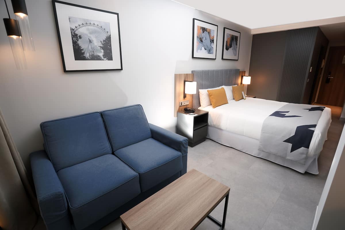 A hotel room with a blue sofa, a wooden coffee table, a bed with white bedding and star patterns, a nightstand with a lamp, and framed pictures on the wall.
