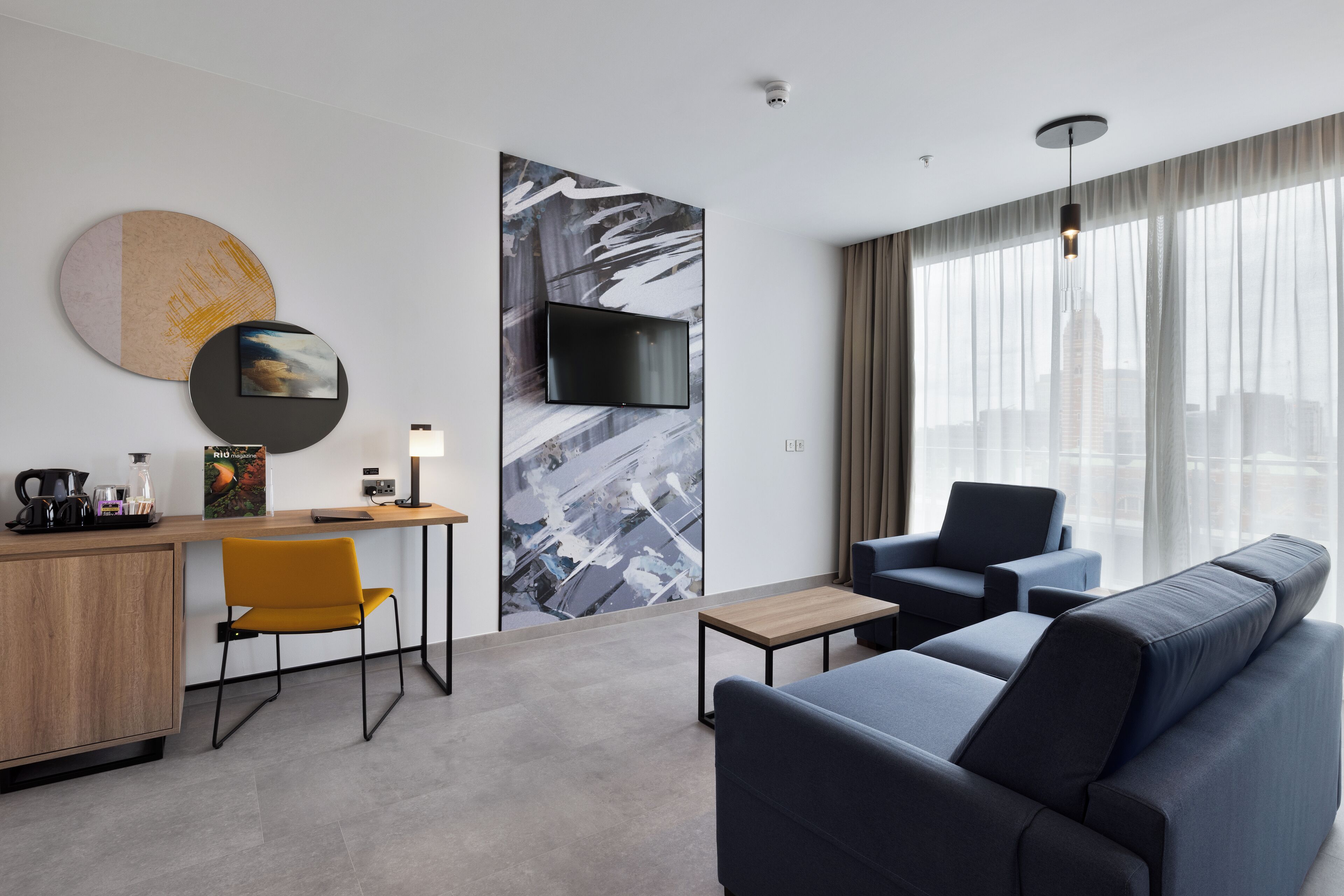 A modern hotel room with a sofa, a desk, a chair, a television, and a large window with a city view.