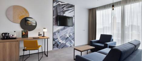 A modern hotel room with a sofa, a desk, a chair, a television, and a large window with a city view.