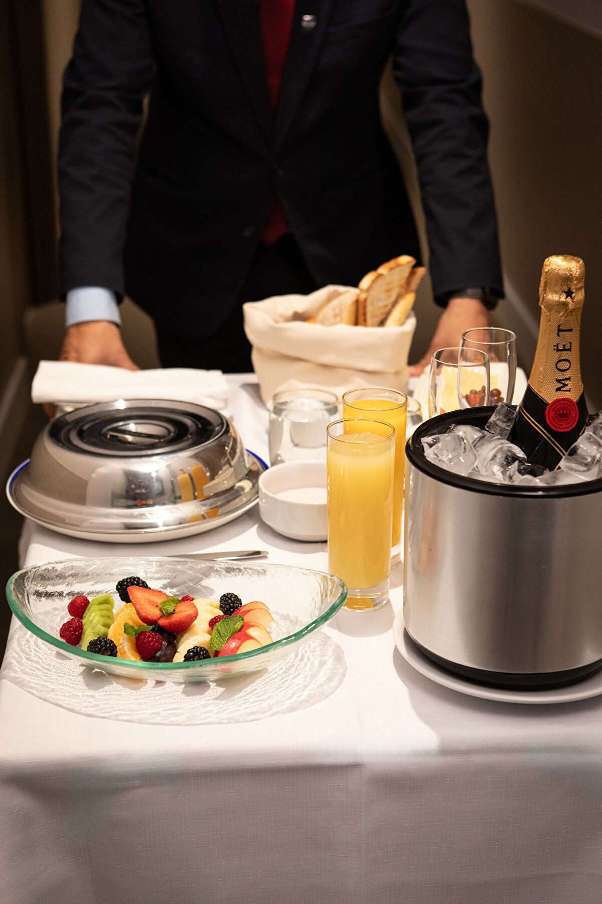 A breakfast tray with a fruit bowl, glasses of orange juice, a bottle of champagne, and a bread basket.