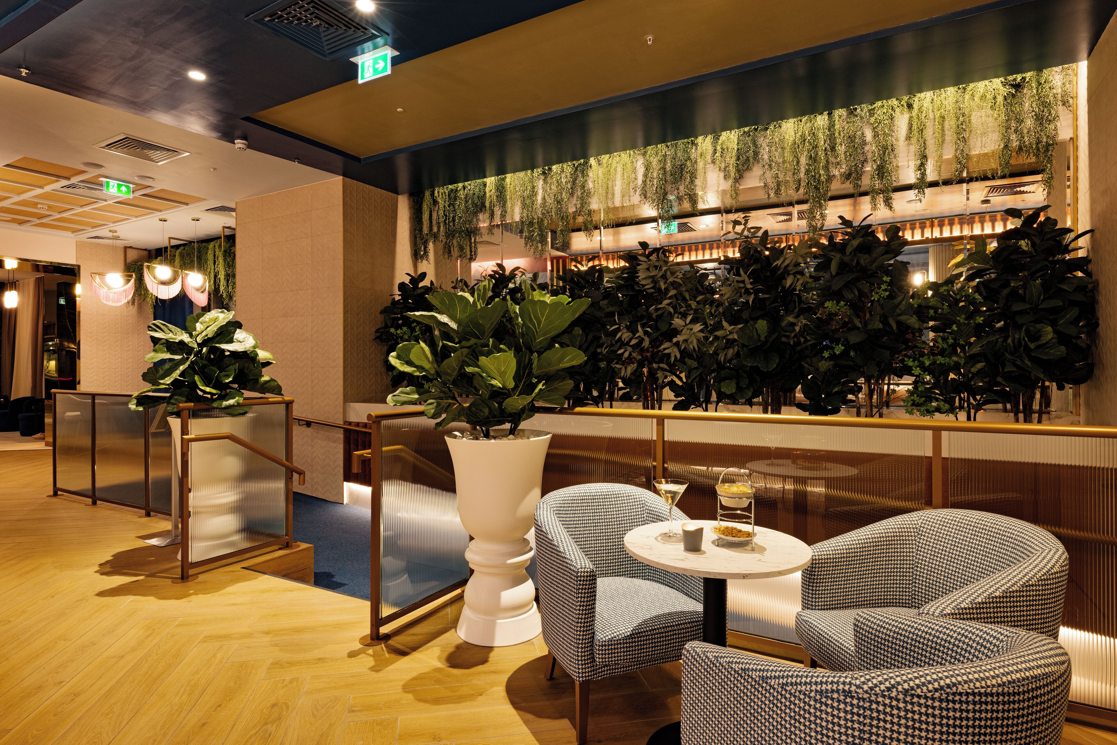 A modern indoor space with wooden flooring, a seating area with checkered chairs, a white pedestal plant, and a wall with hanging greenery.
