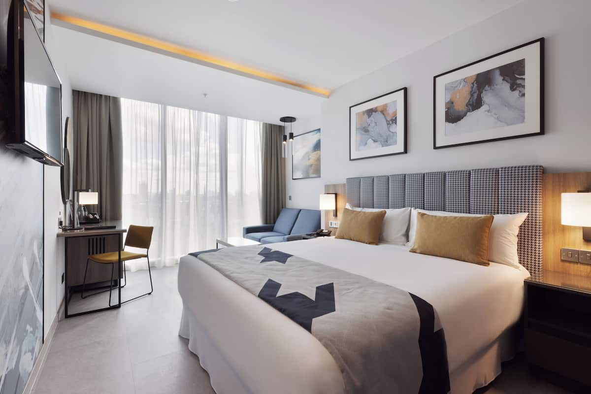 A hotel room with a large bed, a desk with a chair, a TV, and a large window with curtains.