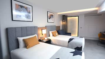 A hotel room with two beds, a desk, and a chair. There are framed pictures on the wall and a window with curtains.