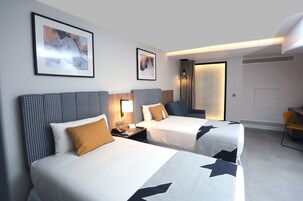 A hotel room with two beds, a desk, and a chair. There are framed pictures on the wall and a window with curtains.