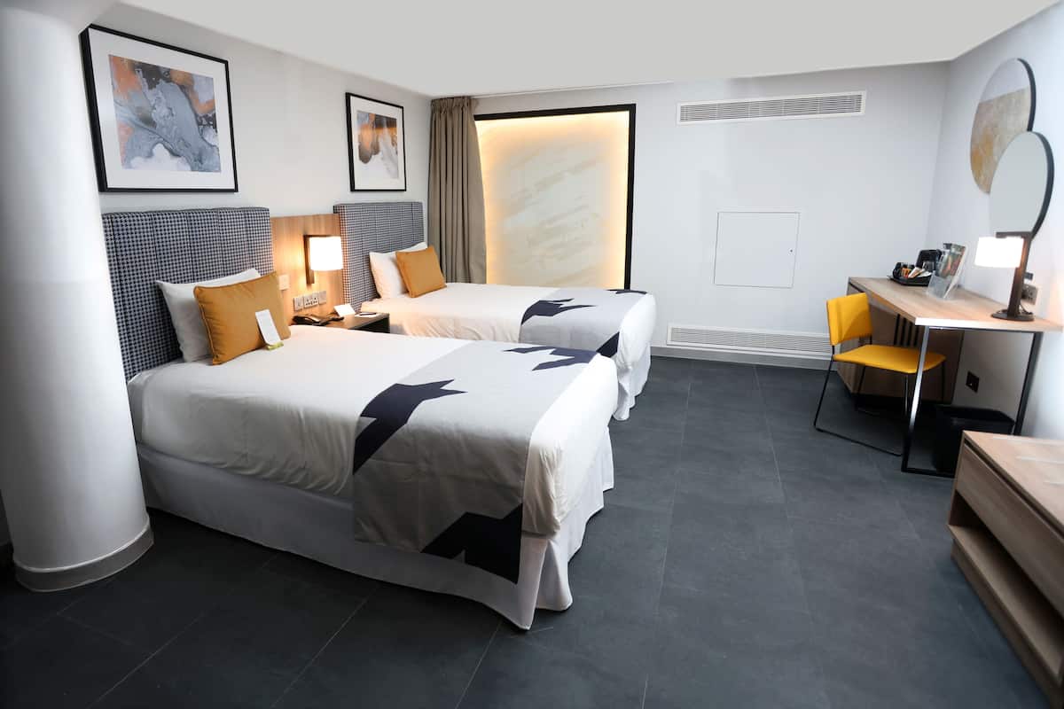 A hotel room with two beds, a desk, a chair, a mirror, and artwork on the walls.