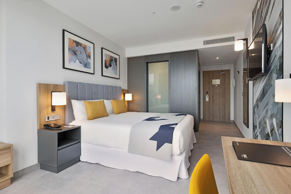 A modern hotel room with a large bed, bedside tables, a desk, and artwork on the walls.