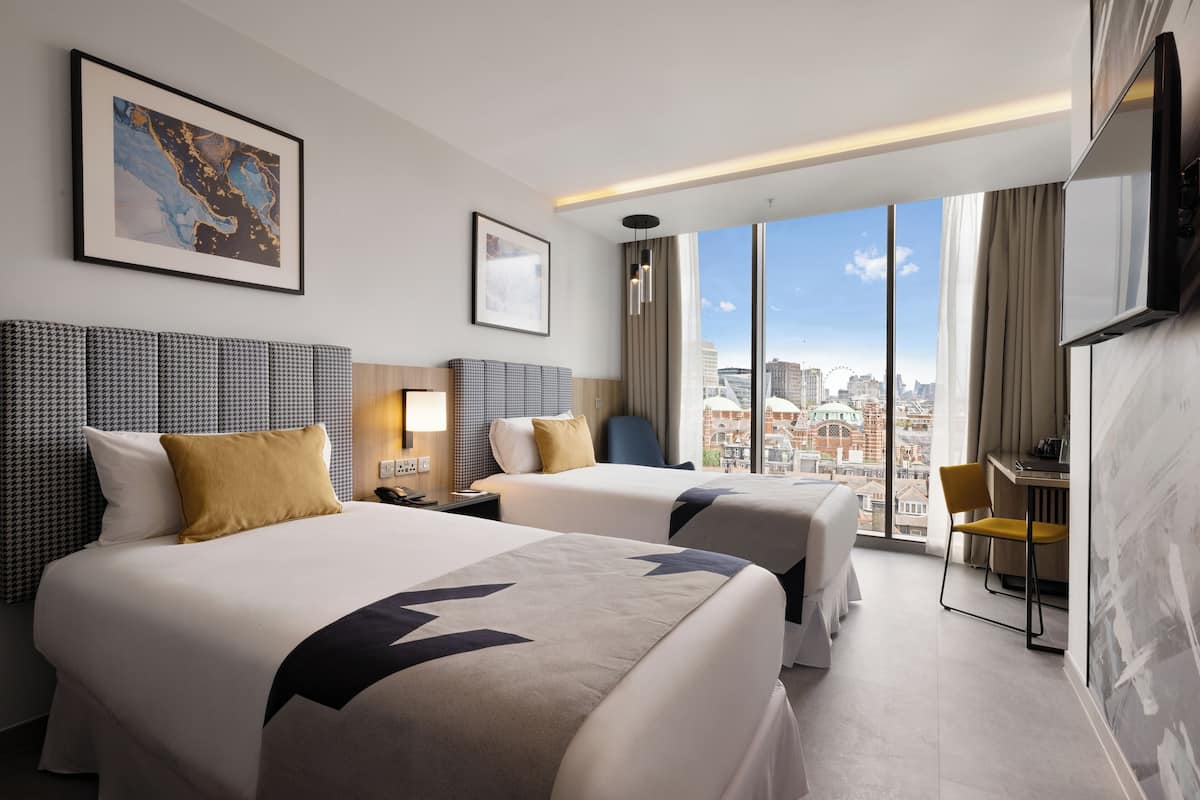 A hotel room with two beds, a desk, and a large window offering a city view.