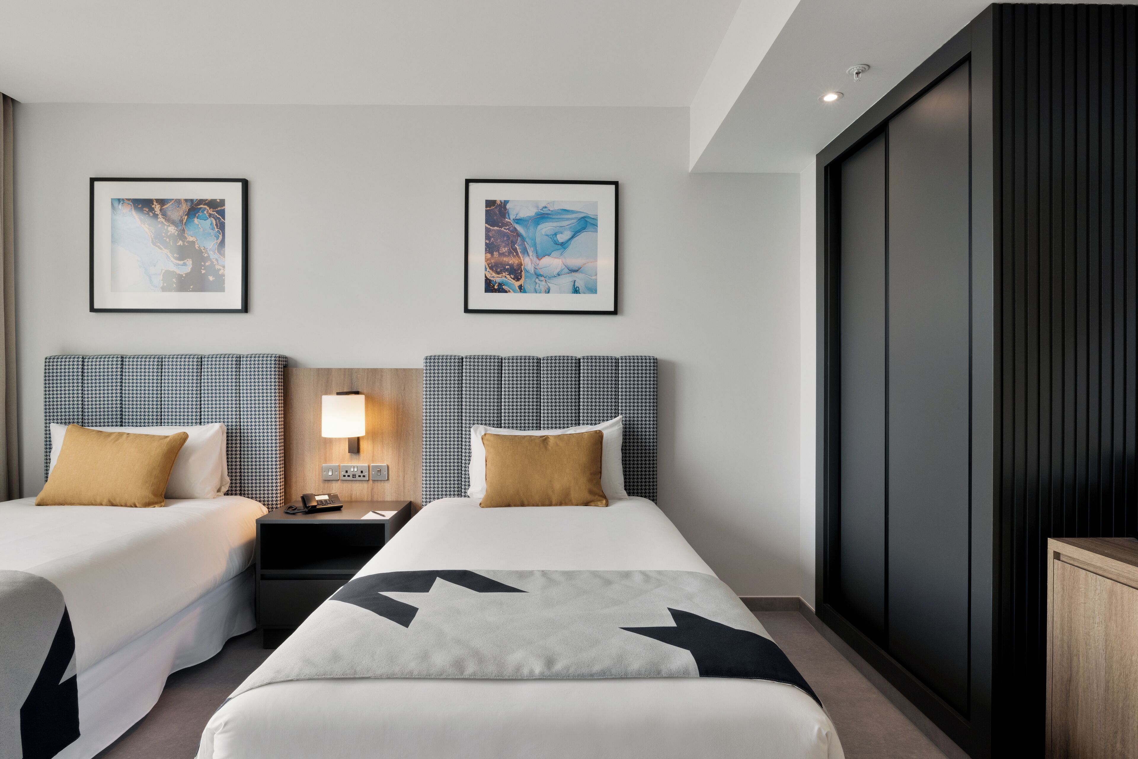 A hotel room with two beds, a nightstand with a lamp, and two framed abstract paintings on the wall.