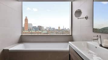 A modern bathroom with a large bathtub, a sink, and a mirror. It features a city view through a large window.