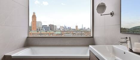 Un baño moderno con una bañera grande, un lavamanos y un espejo. Tiene vistas a la ciudad a través de un amplio ventanal.