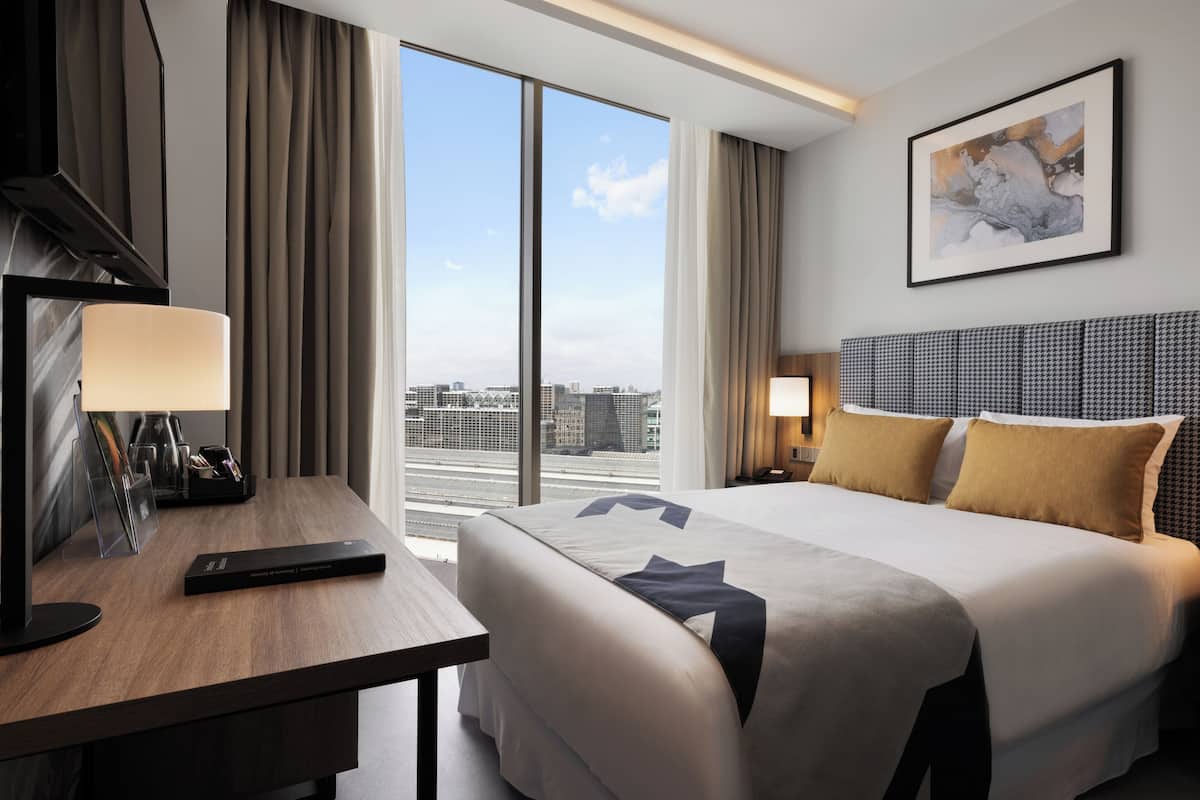 A hotel room with a large bed, a desk with a lamp, a city view through large windows, and a framed artwork on the wall.