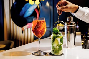 A bar setting with a cocktail glass, a glass with a mint garnish, and a cocktail shaker on a white table.
