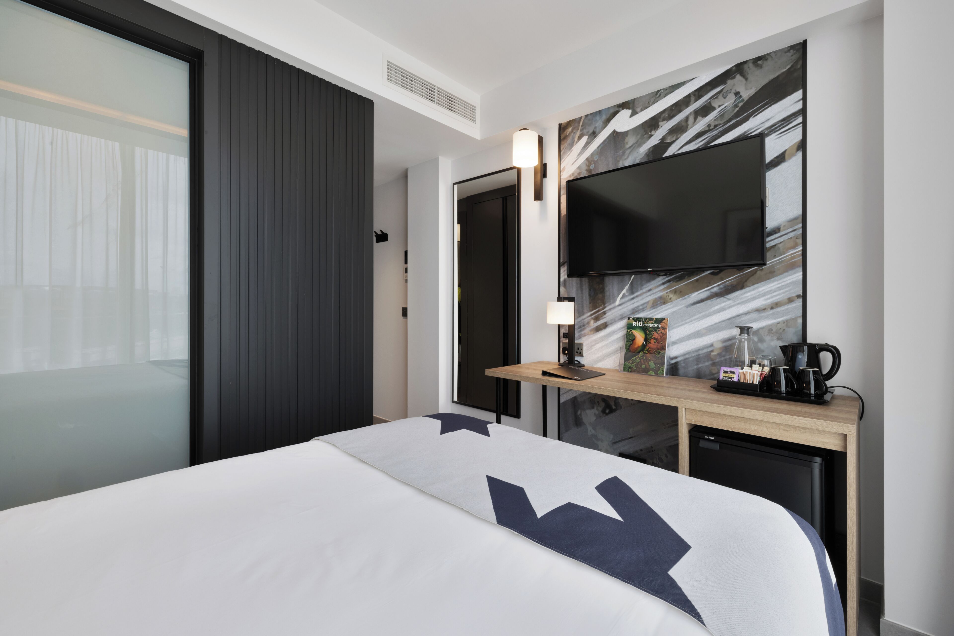 Ett modernt hotellrum med en stor säng, en platt-TV som är monterad på väggen, en liten kaffebryggare och ett skrivbord med en lampa.