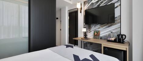 Una habitación de hotel moderna con una cama grande, un televisor de pantalla plana en la pared, una cafetera pequeña y un escritorio con una lámpara.