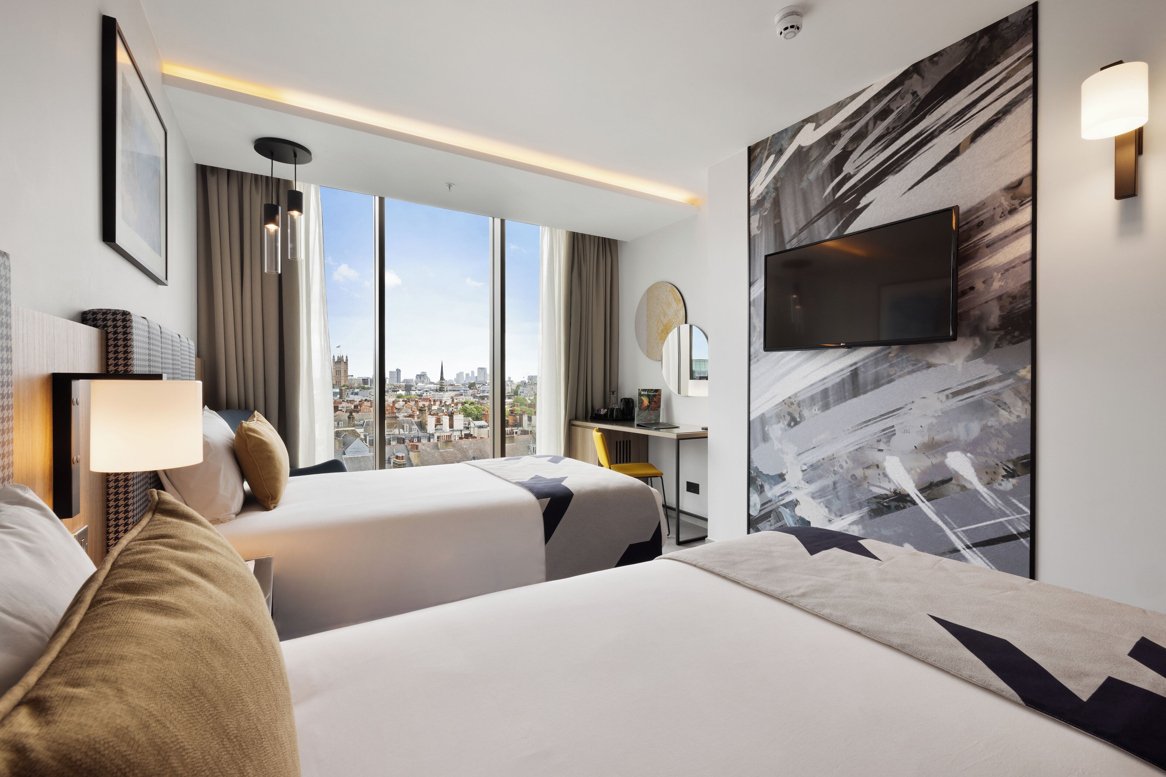 A hotel room with two beds, a desk, a chair, a TV, and a large window with a city view.