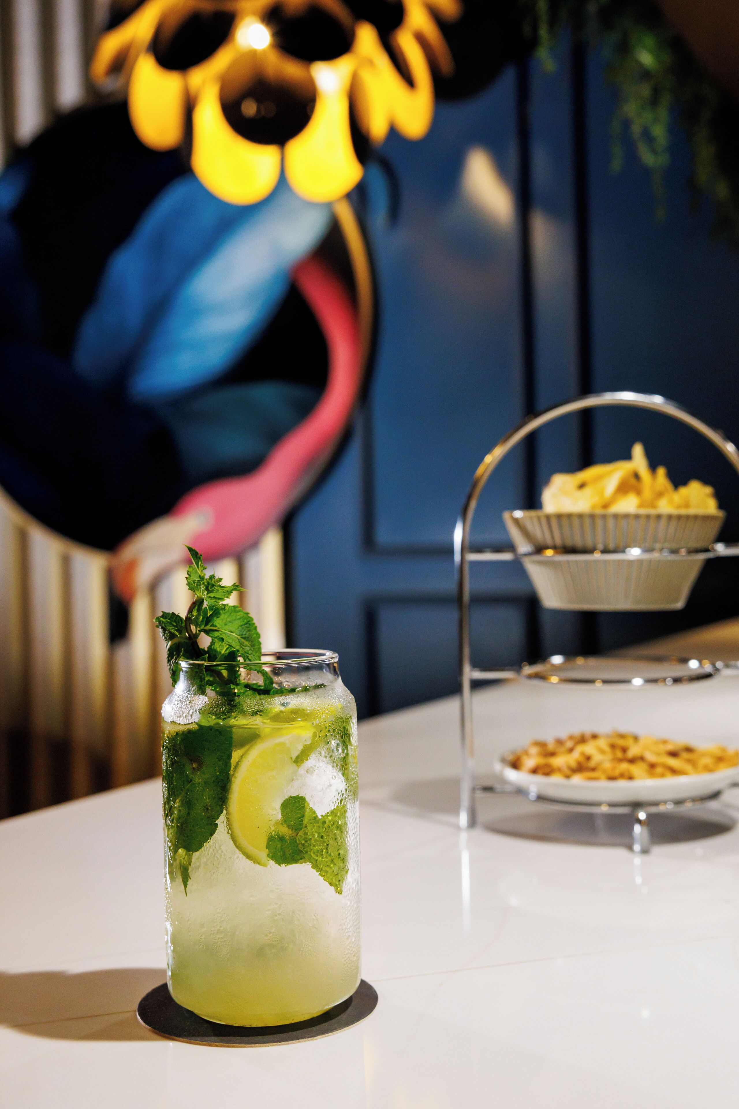 Et glas isdrikk med mynte og lime, en tallerken med chips, og en dekorativ flamingo i baggrunden.