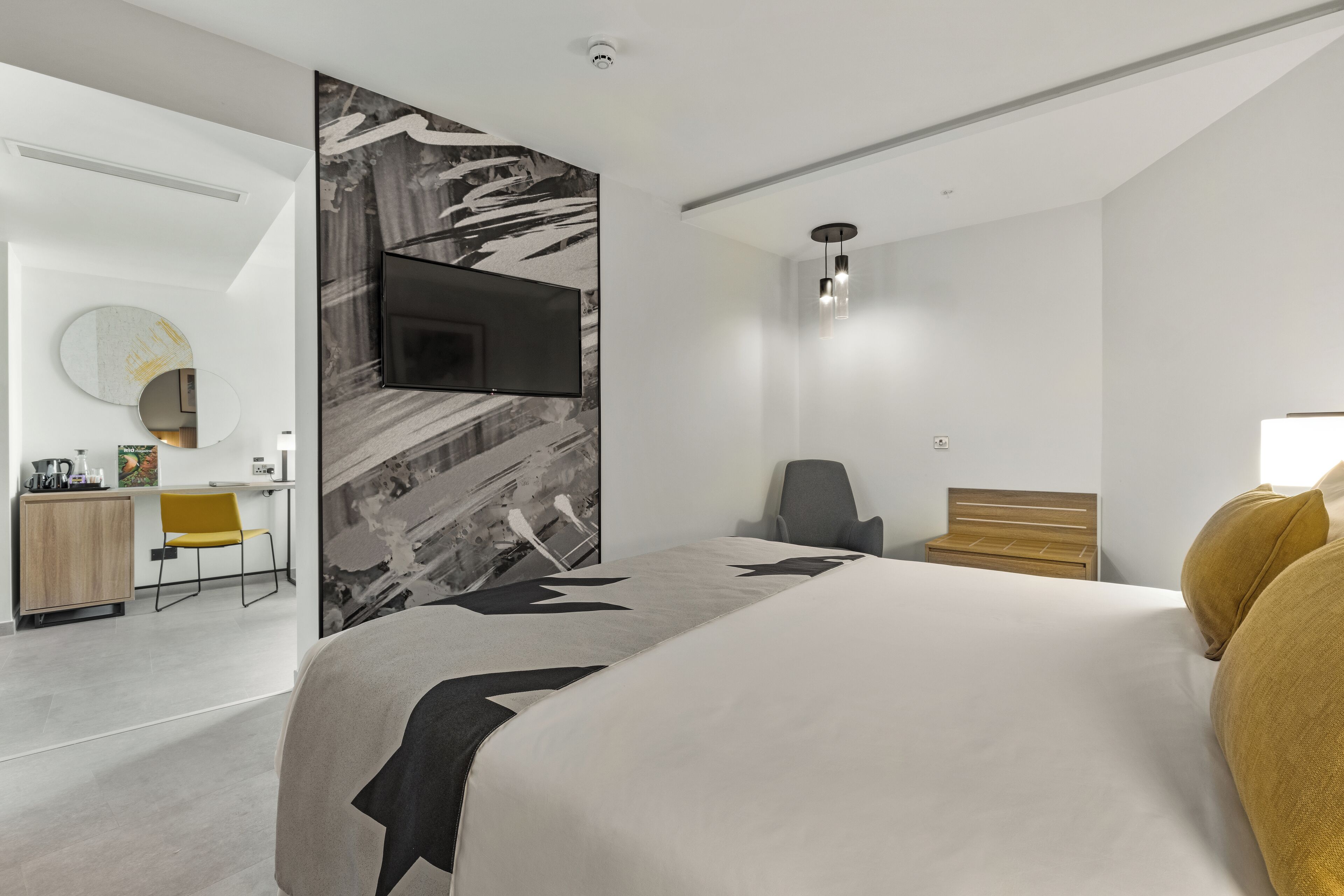 A modern hotel room with a large bed, a flat-screen TV mounted on the wall, a desk with a chair, and a mirror.