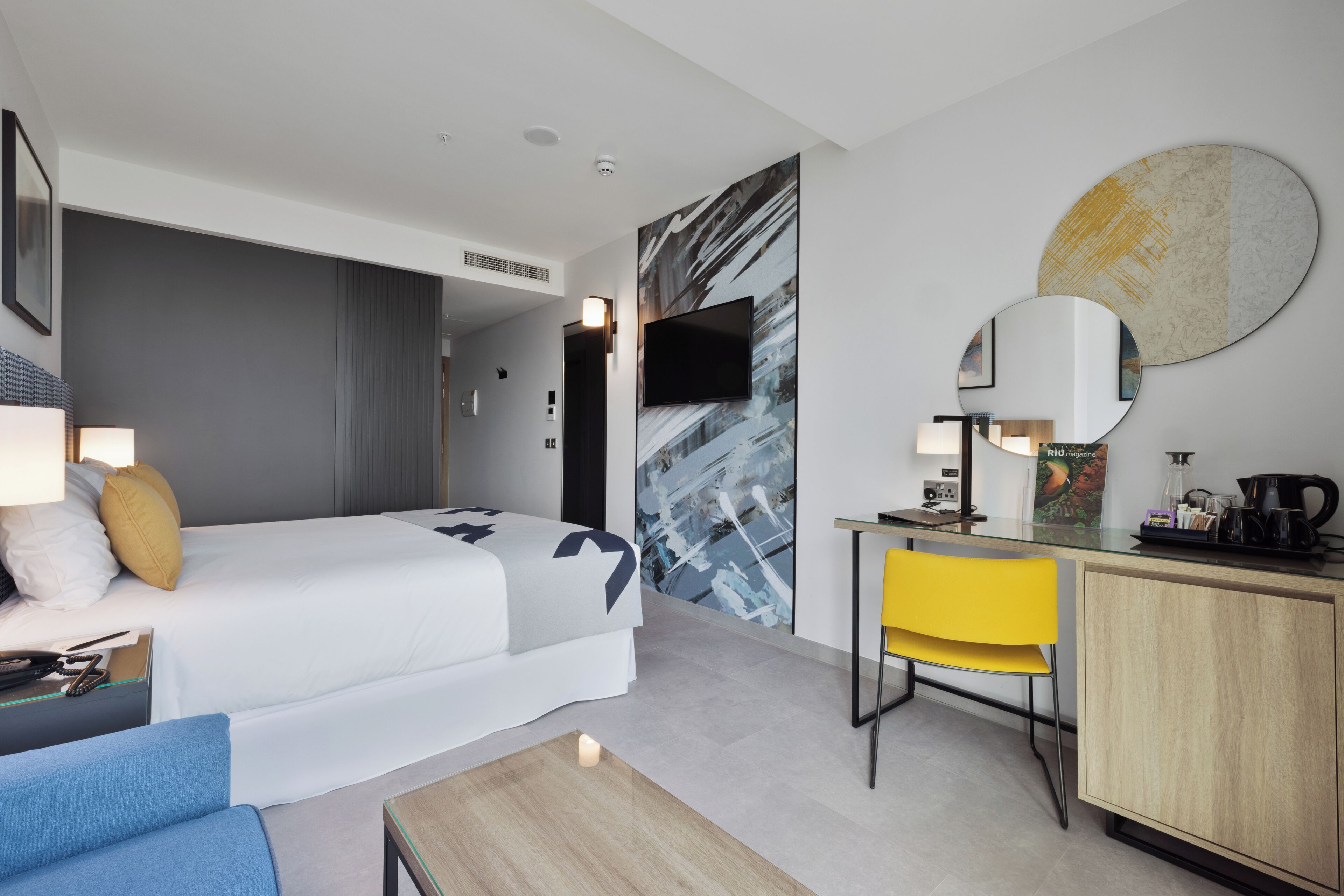 A modern hotel room with a bed, a desk, a chair, and a small kitchenette.