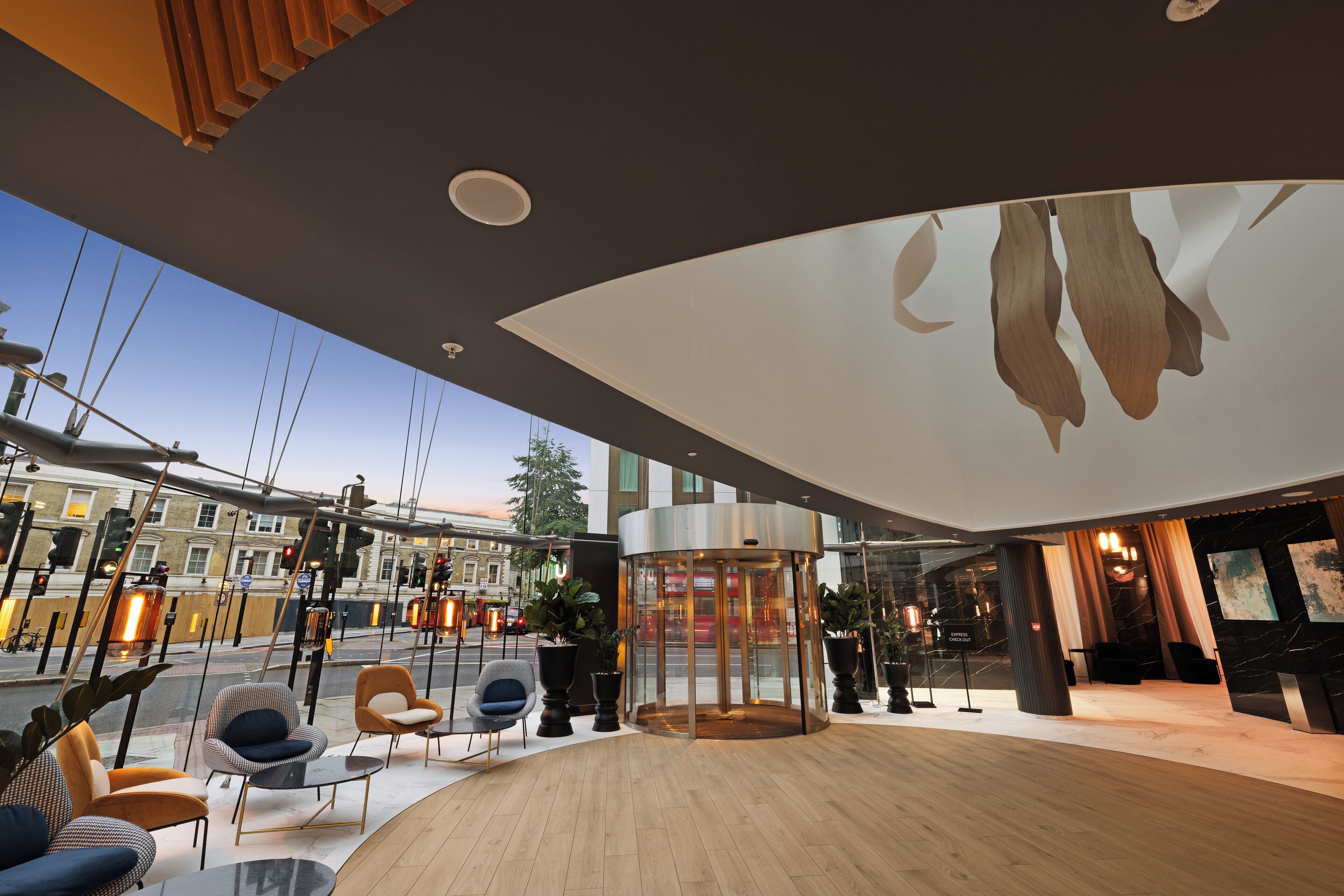A modern hotel lobby with a central rotating door, seating area with chairs and tables, and large windows offering a view of the street.