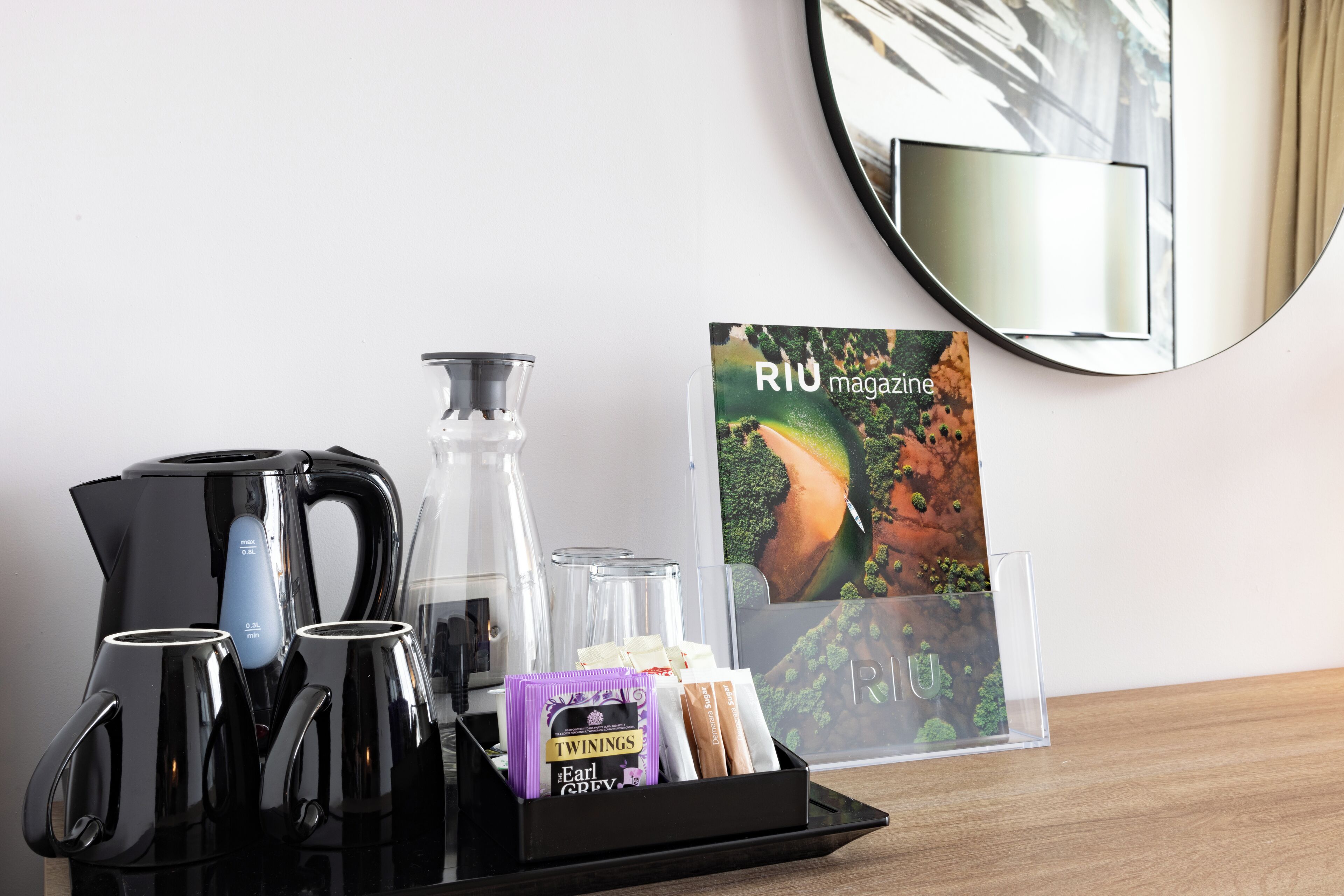 A coffee station with a black coffee maker, two glass carafes, a glass pitcher, and a tray with coffee packets and a magazine titled "RIU magazin".