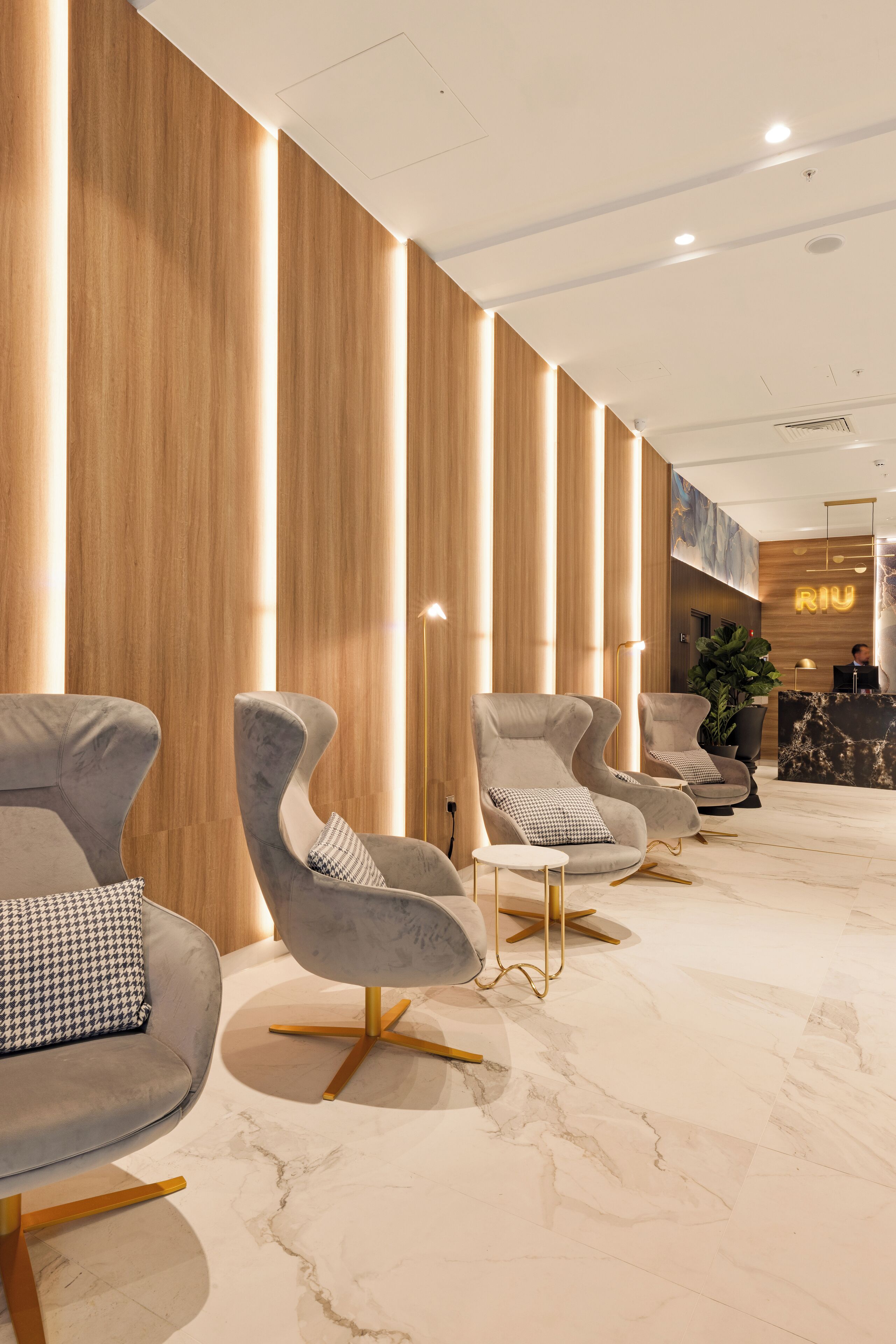 A modern lobby with a row of grey upholstered chairs with gold legs, a marble floor, and wooden paneling on the walls.