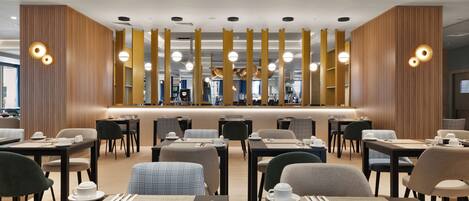 A modern hotel dining area with neatly arranged tables and chairs, a central counter, and ambient lighting.