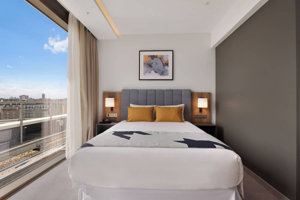 A hotel room with a large bed, two bedside tables with lamps, a framed artwork on the wall, and a city view through the window.