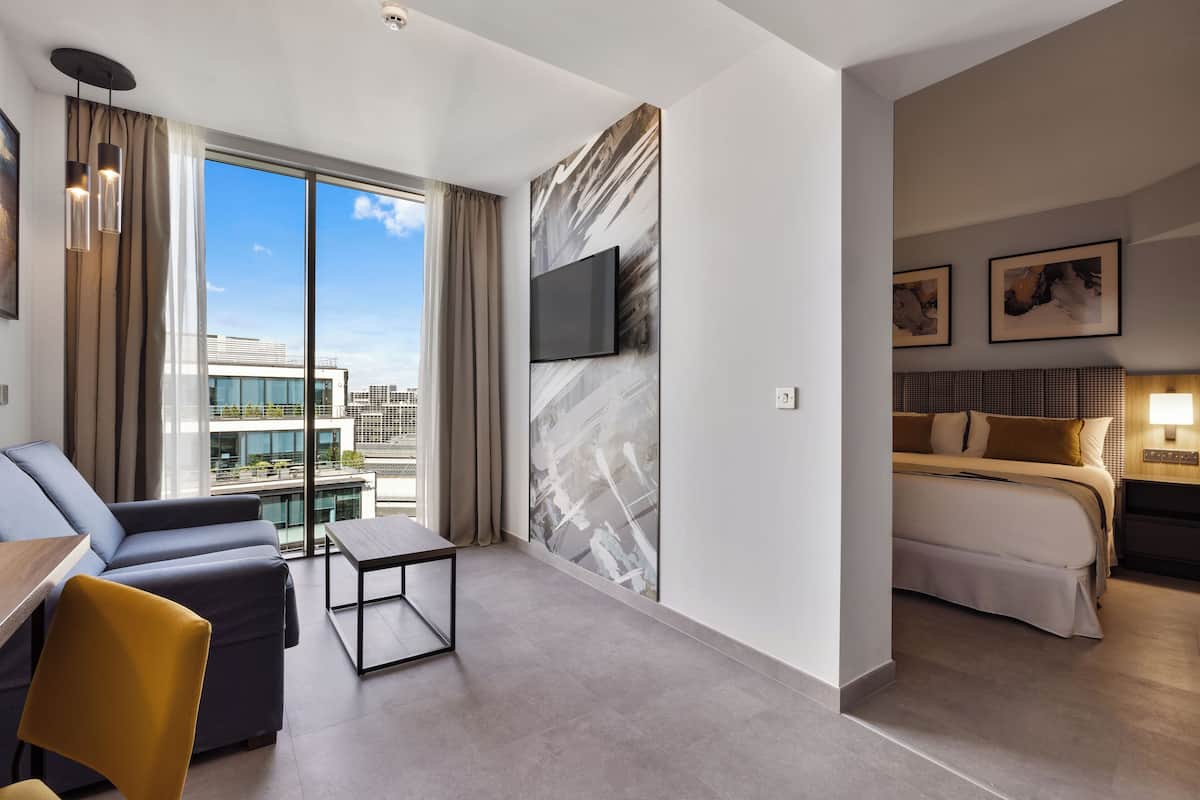 A modern hotel room with a large window, a flat-screen TV, a bed, a sofa, and a small table.